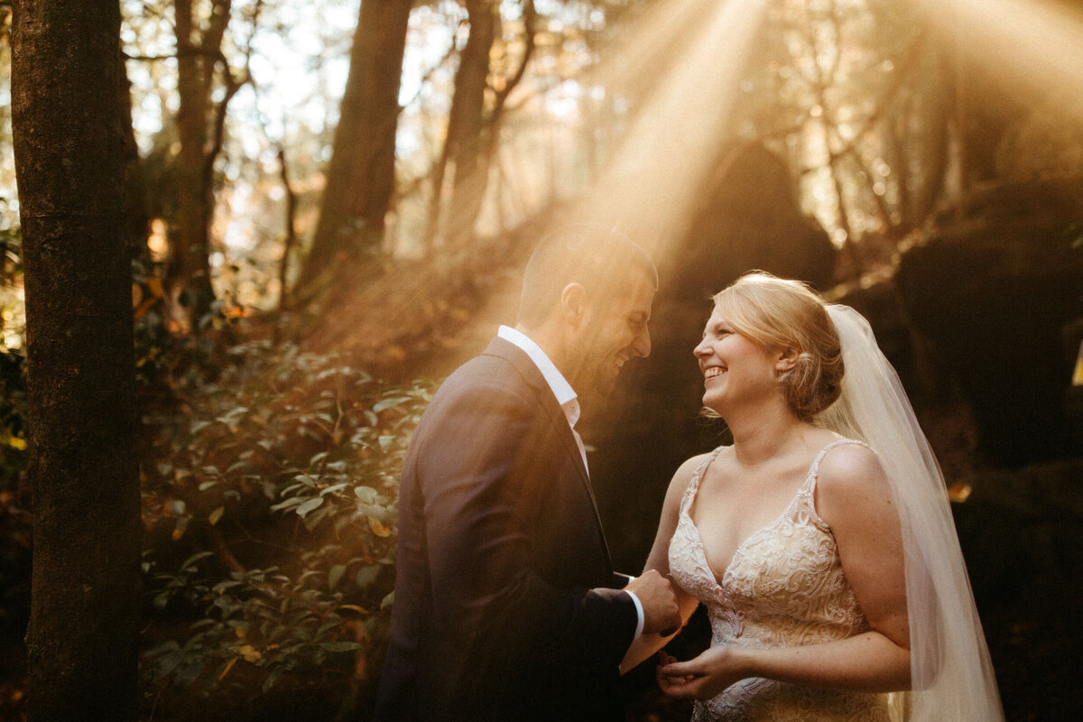 Bride and groom laughing together in the woods with golden sun rays shining around them