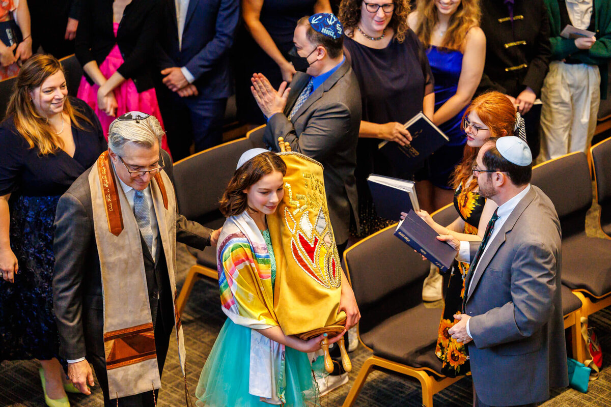 A teen girl in a blue dress and rainbow tallit carried the torah to the bimah followed by a rabbi allowing Bellevue Bar and Bat Mitzvah Photography
