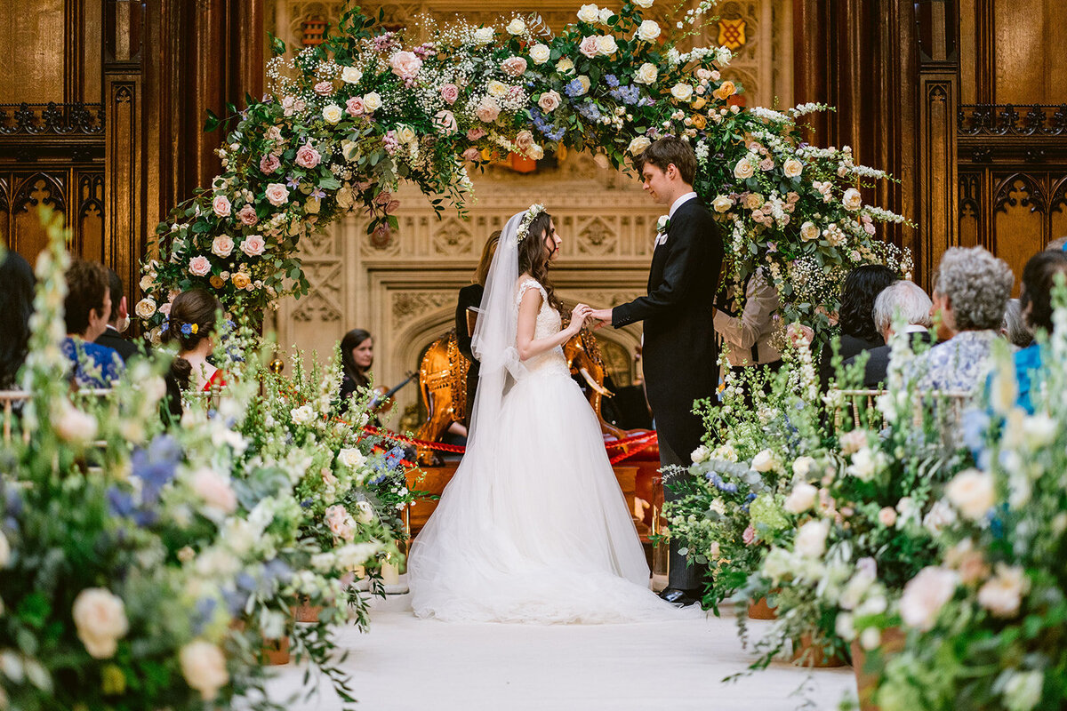 bride and groom exchanging rings in the great hall under a flower arch at at allerton castle