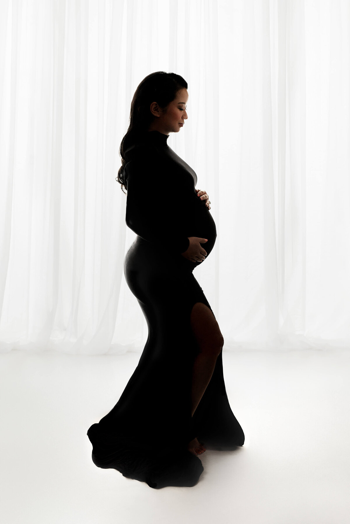 pregnant mother in a black dress standing on front of a white curtain at a maternity photography session with a photographer at a northern virginia photography studio