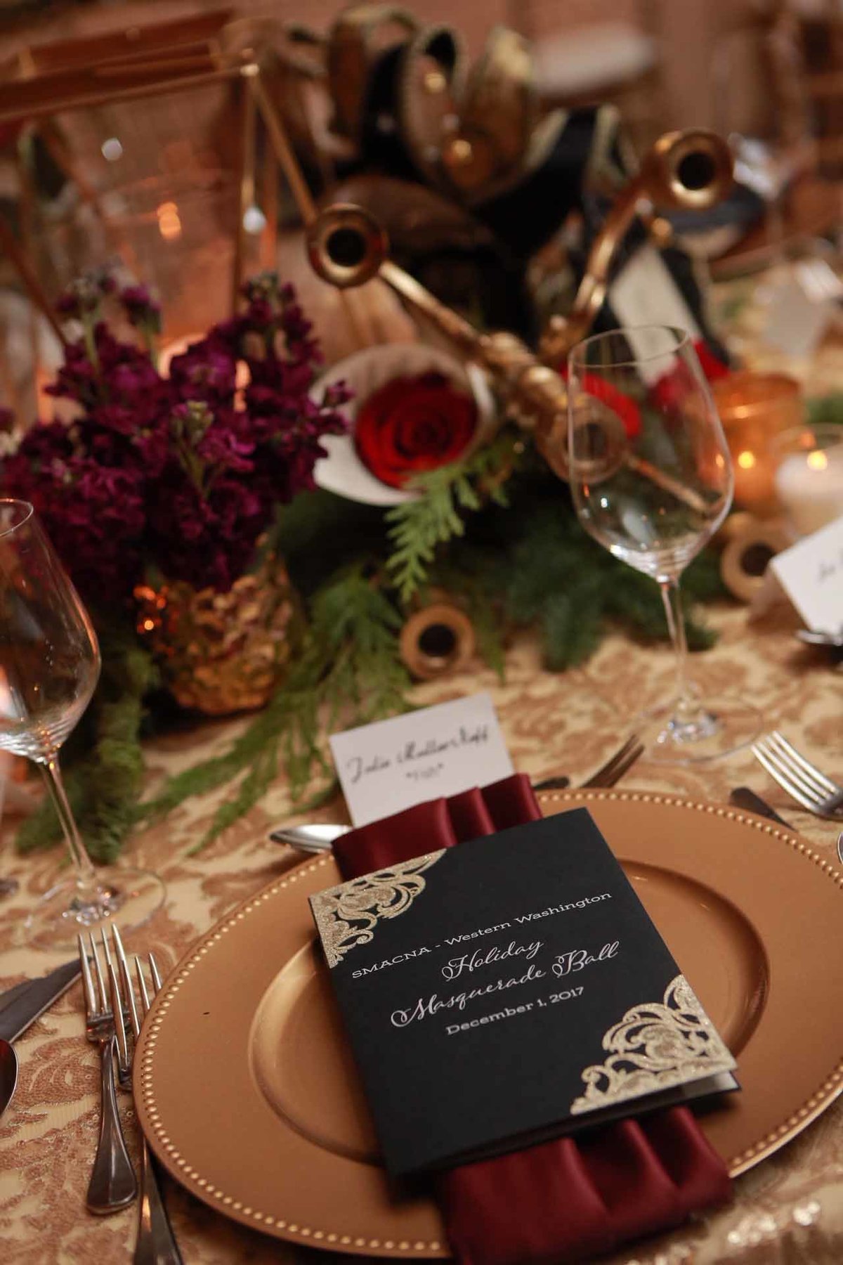 holiday party place setting with red napkin, black menu card on gold charger plate and evergreen garland runner