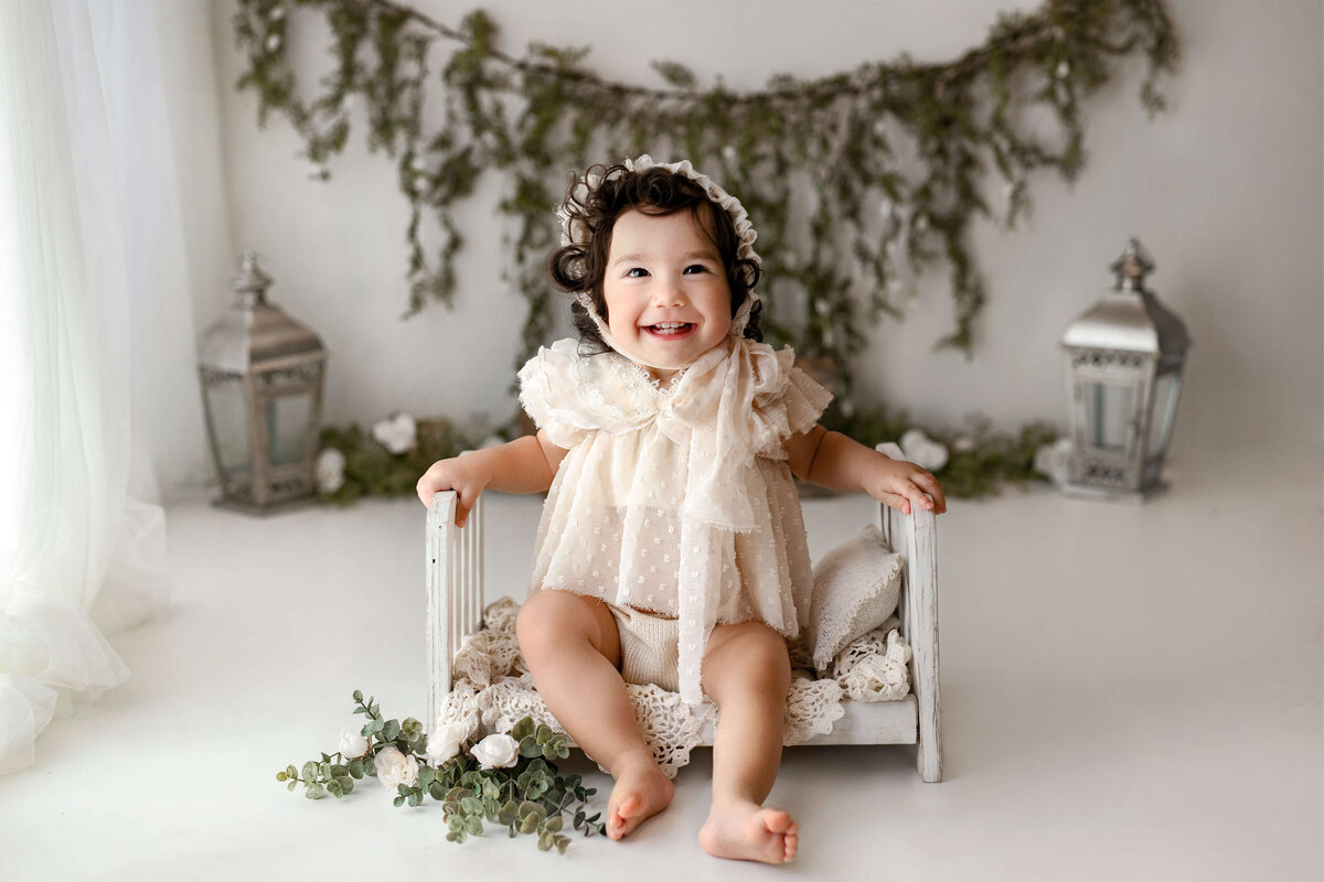 toddler wearing a cream outfit and bonnet sitting on a white wooden bed with greenery and a white background