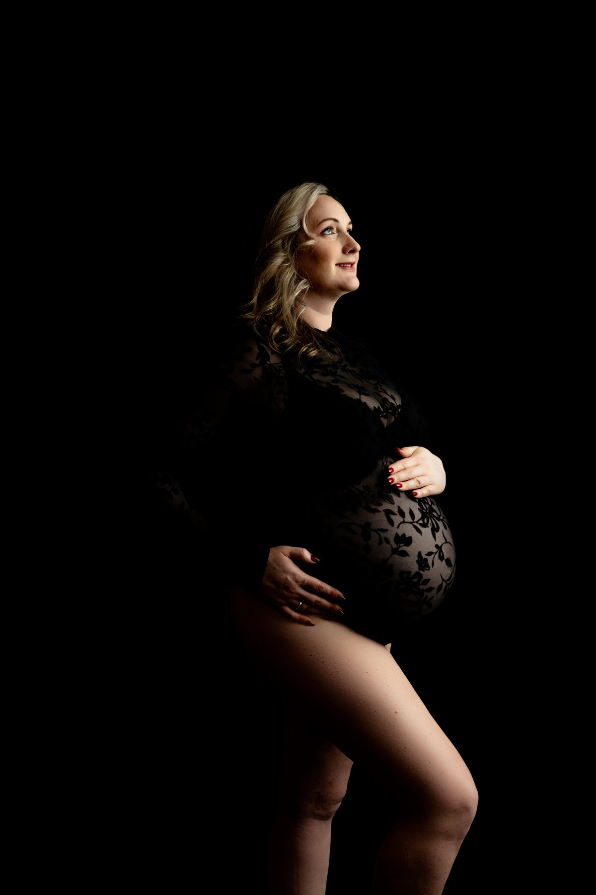A happy mother to be in a black lace one-piece holds her bump while standing in a New Jersey Maternity Photographer studio