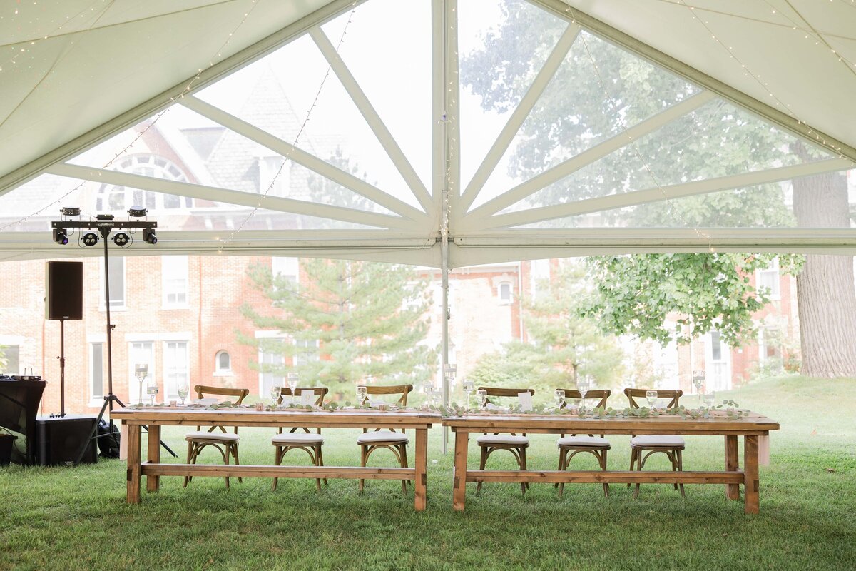 Elegant outdoor wedding setup under a large tent with wooden tables, chairs, and place settings in an Iowa park. Trees and buildings are visible in the background.