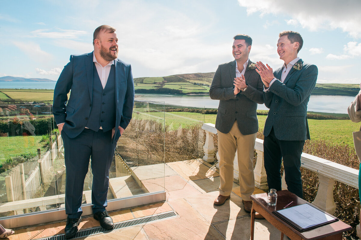 two grooms clapping on a sunny terrace laughing and looking at guest