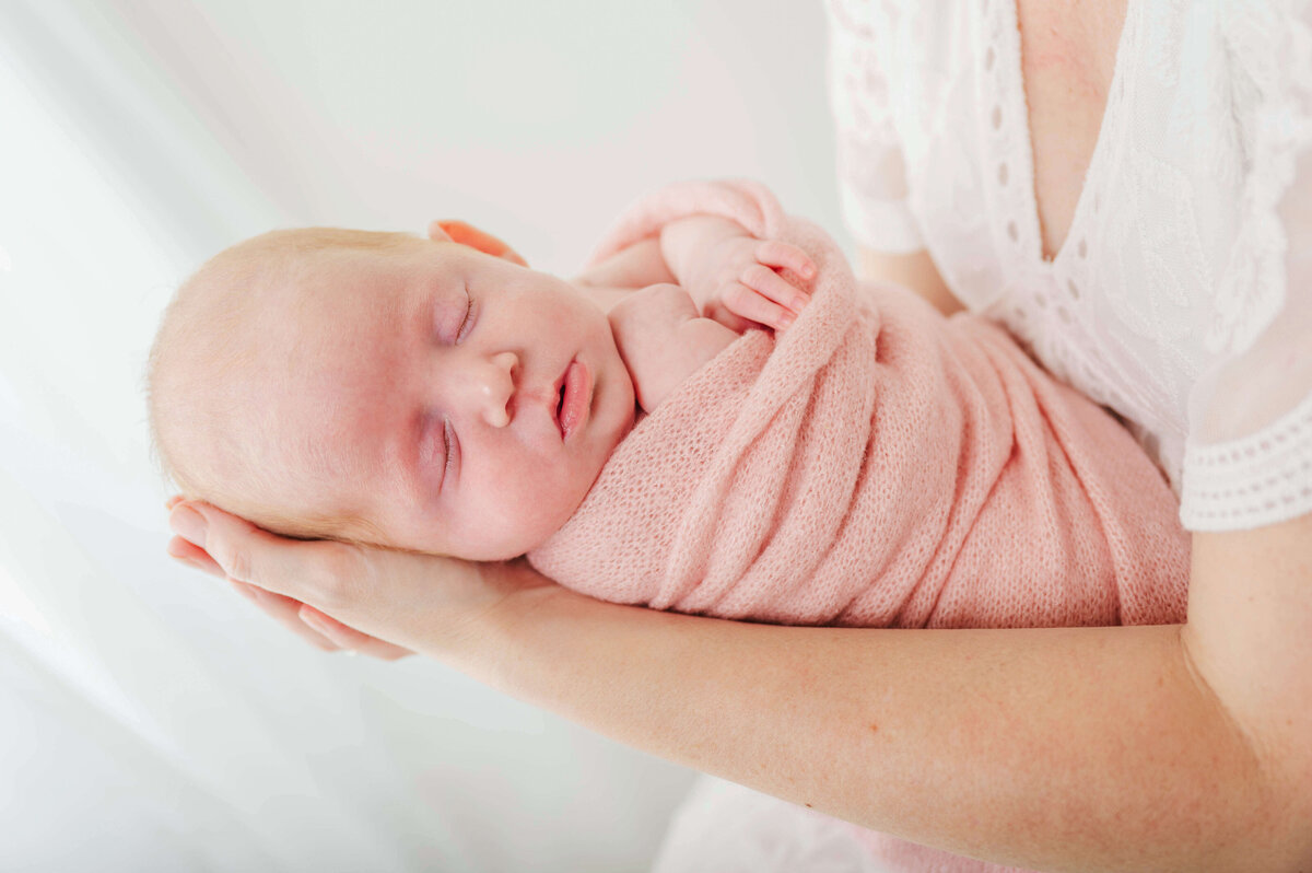 A newborn wrapped in pink blanket and laying in mom's arms in front of a window with a white sheer curtain.