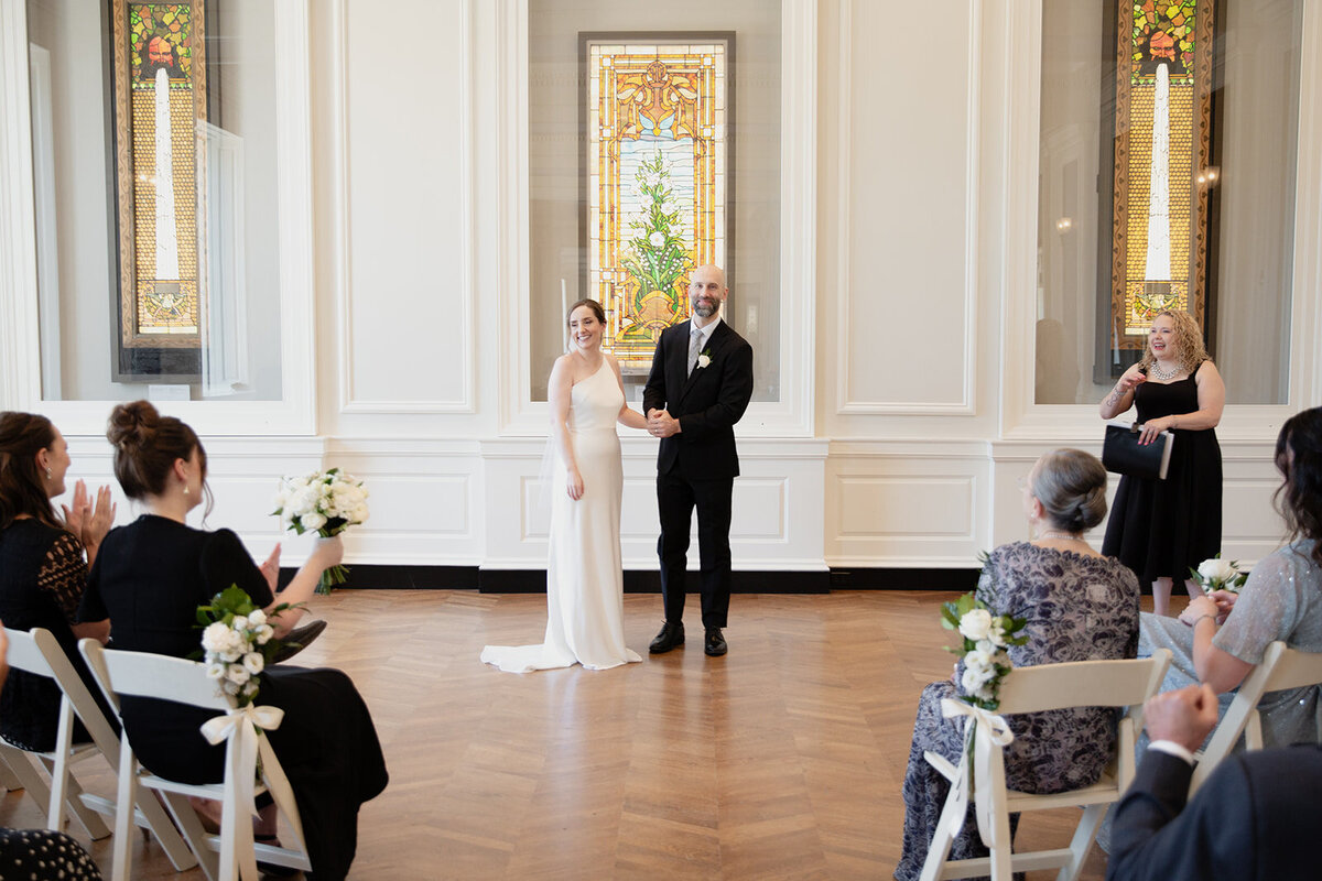 The couple looks gratefully at their guests as they hold hands, adn the guests applaud.