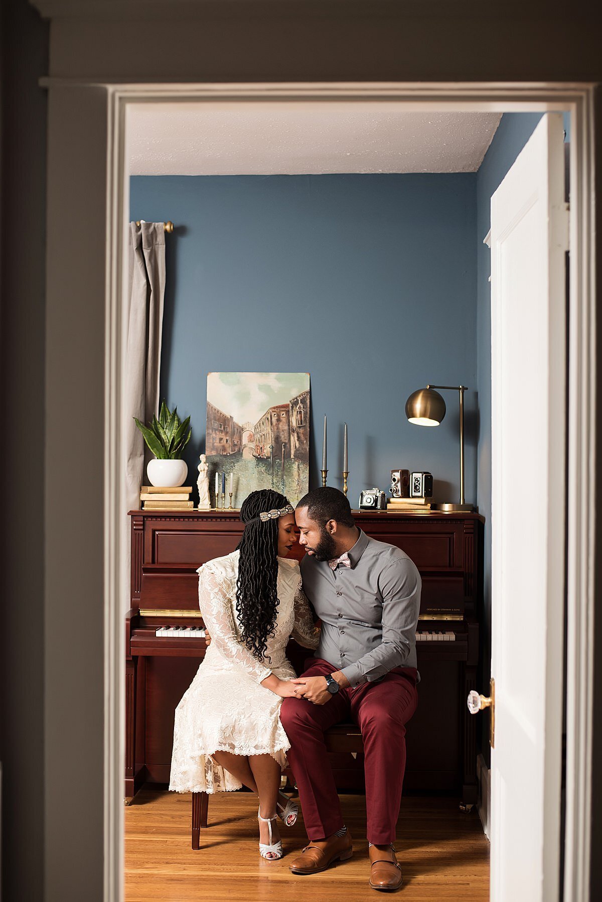 The bride and groom sit in a room with blue walls on a piano bench with an upright piano behind them. The bride is wearing a lace headband and a long sleeved lace fit and flare wedding dress that comes to her knees with t-strap heels. The  bride and groom are touching foreheads and holding hands. The groom is wearing a gray button down shirt with maroon pants and brown shoes. The photo is being taken through an open door with a crystal door knob.