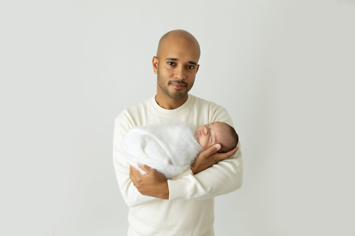 A father in a white shirt cradles his sleeping newborn baby in his arms