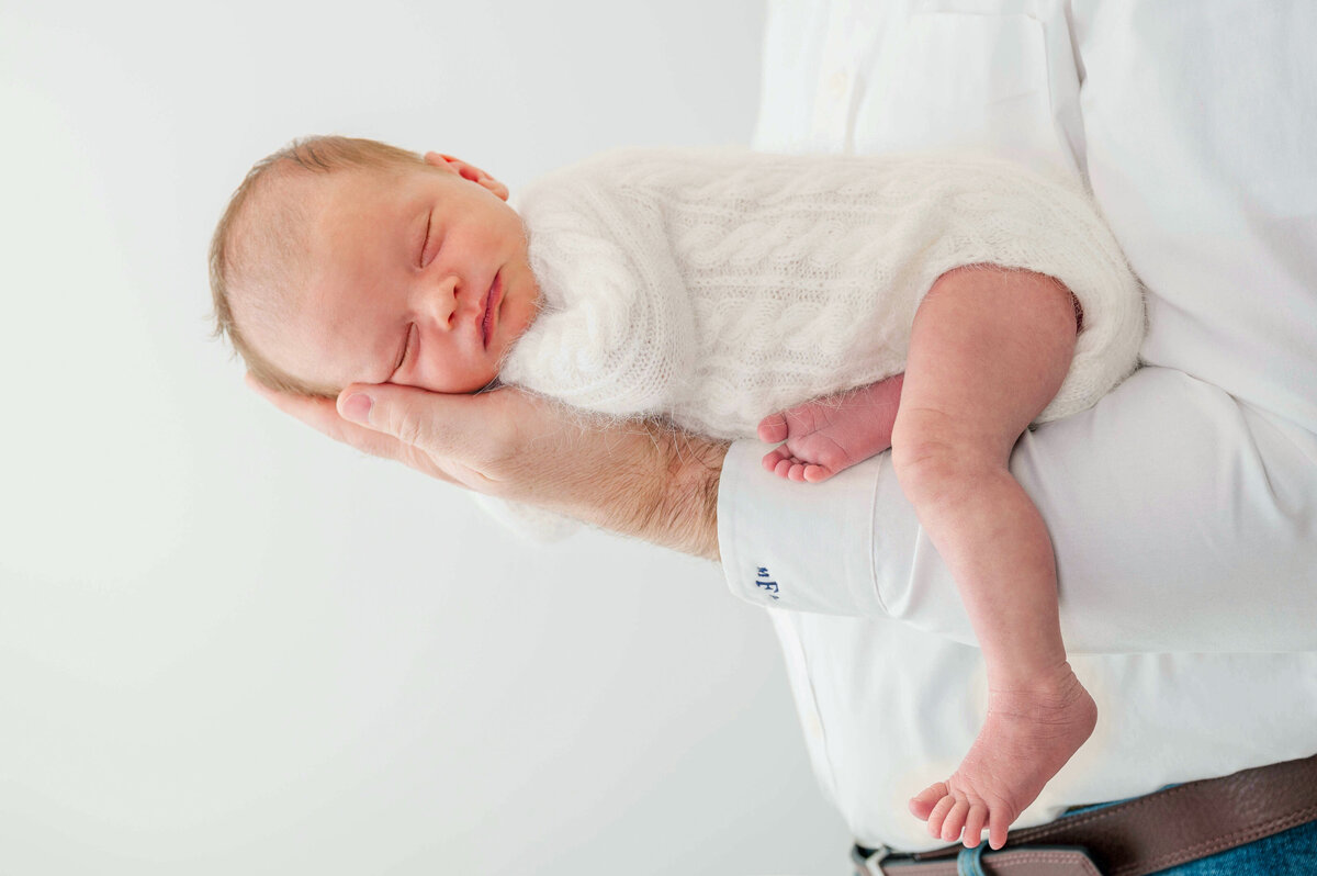 A newborn baby girl in a white sweater sleeping on stomach on dad's arm, against a white backdrop.