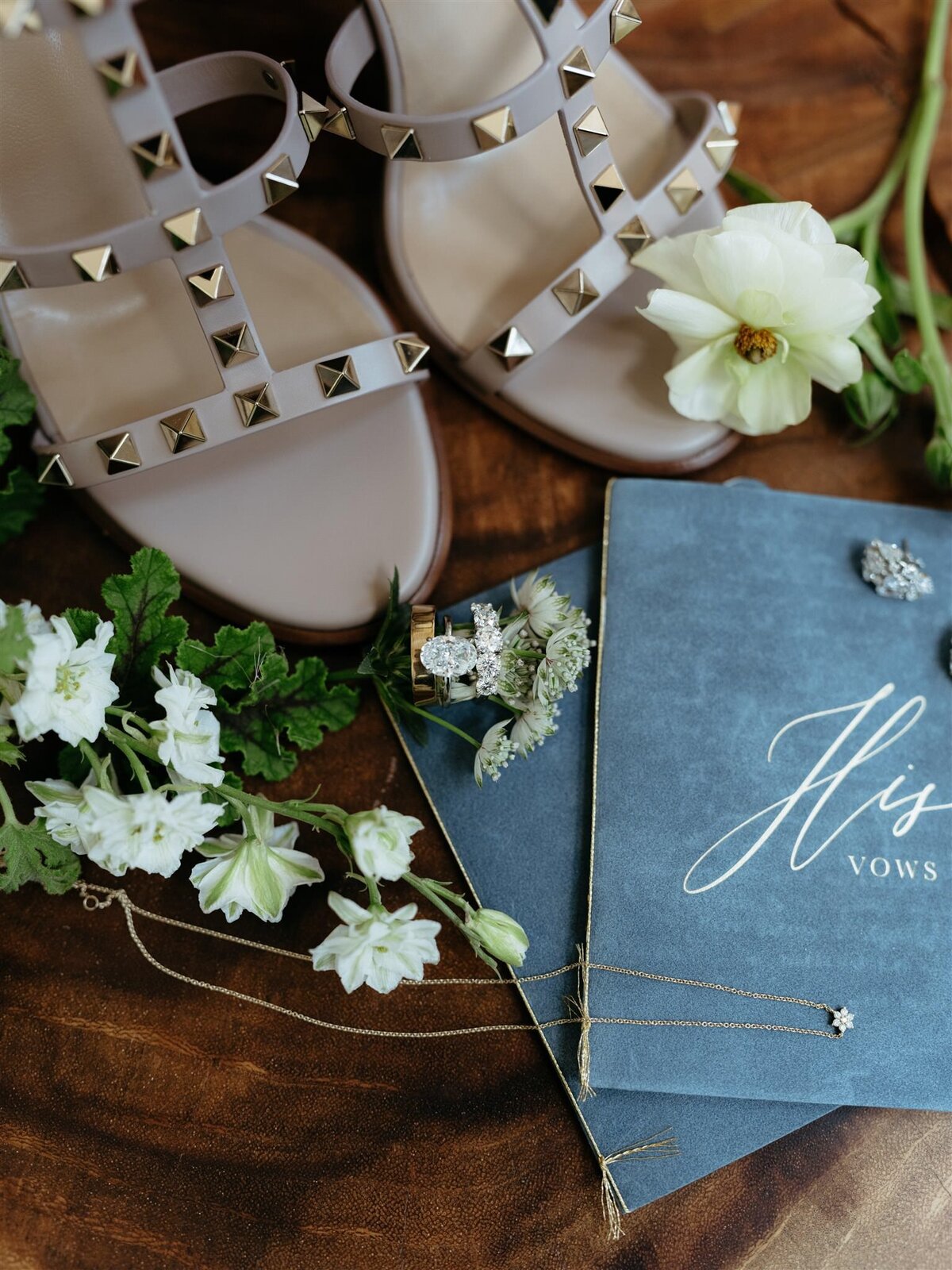 Detail photo of bride's gold-studded shoes, white floral accents, dainty gold necklace, and little blue book of vows.