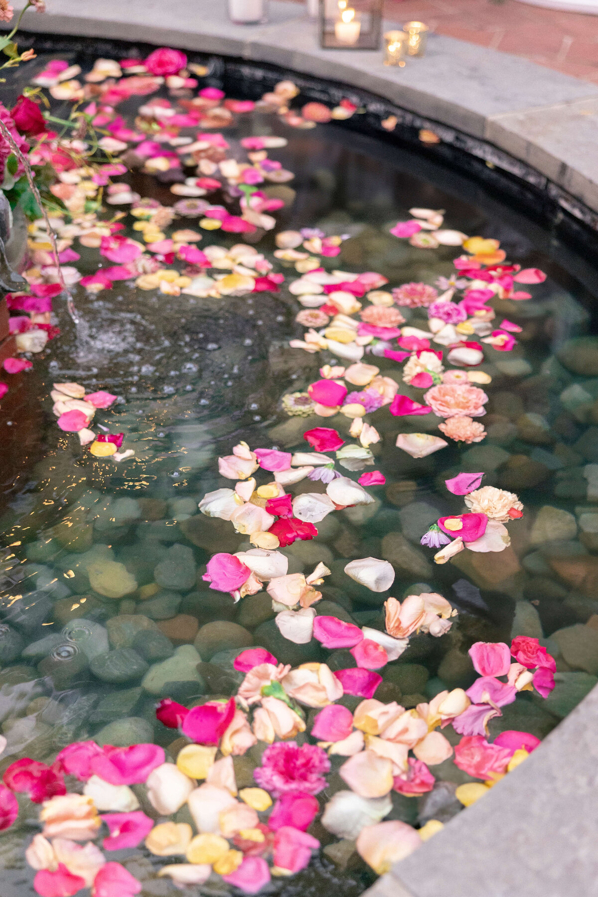 A small pond with clear water, filled with floating rose petals in shades of pink, red, yellow, and white. The pond is surrounded by a stone border, and a few candles are visible in the background.