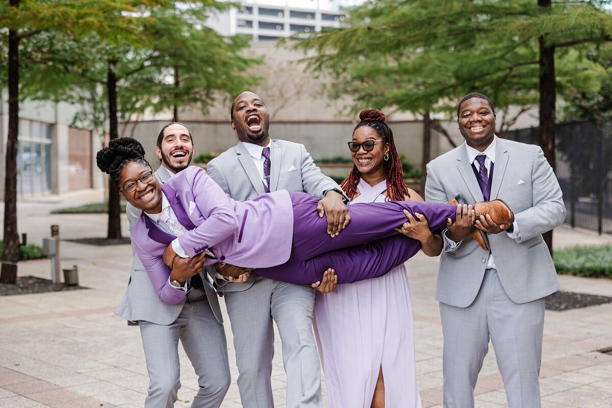 A portrait of a bride being playfully held up by her portion of the wedding party on her wedding day at The Westin Dallas Downtown in Dallas, Texas. The bride is wearing a purple suit with a floral tie. Her wedding party is wearing either grey suits with purple ties or a lavender dress.