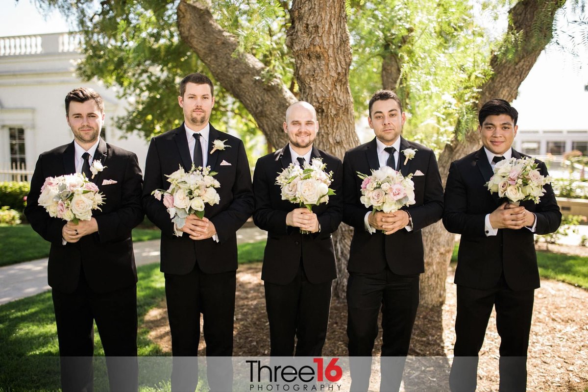 Fun photo of Groomsmen posing holding the Bridesmaid's bouquets