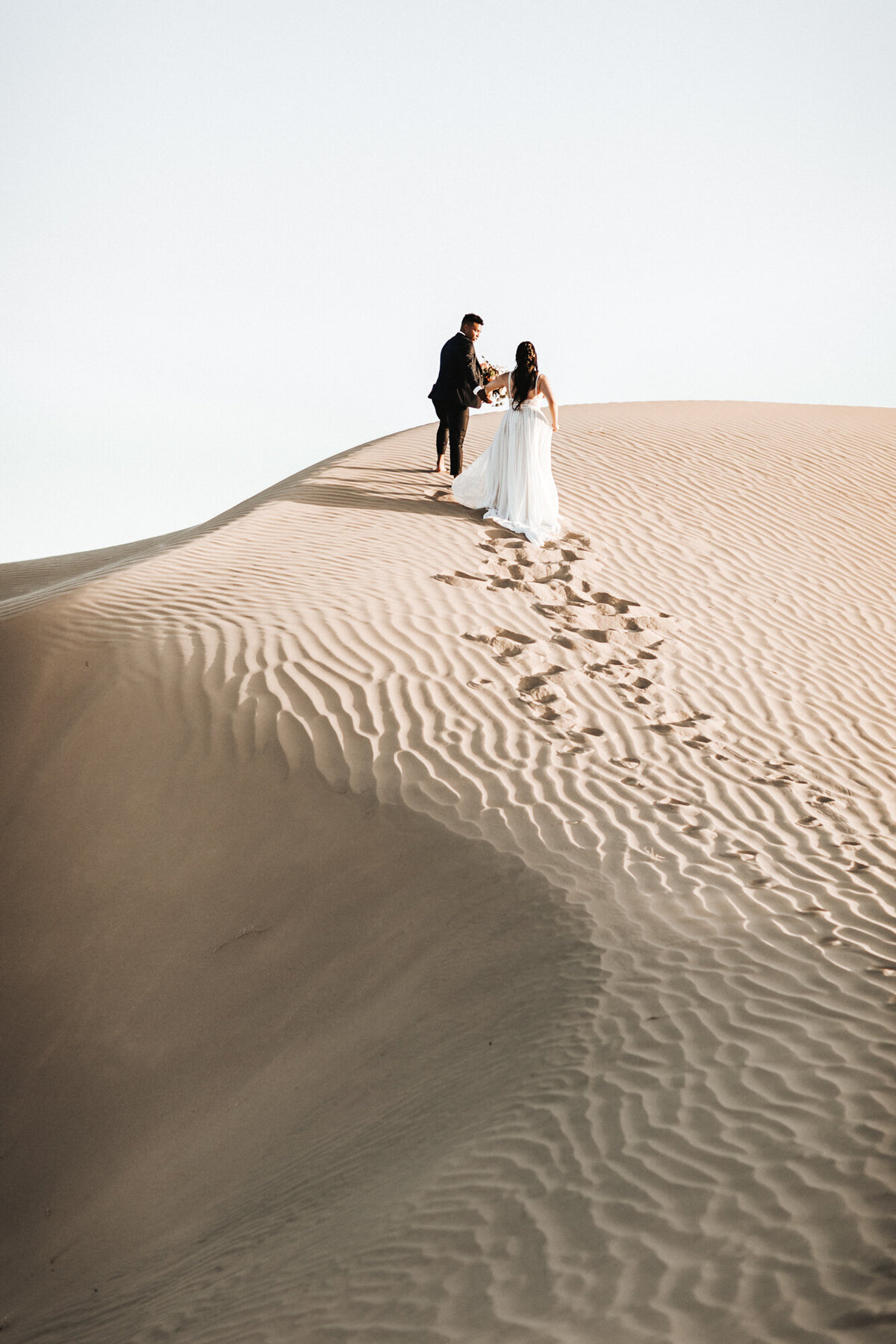 Couple wearing wedding attire climbing a sand dune.