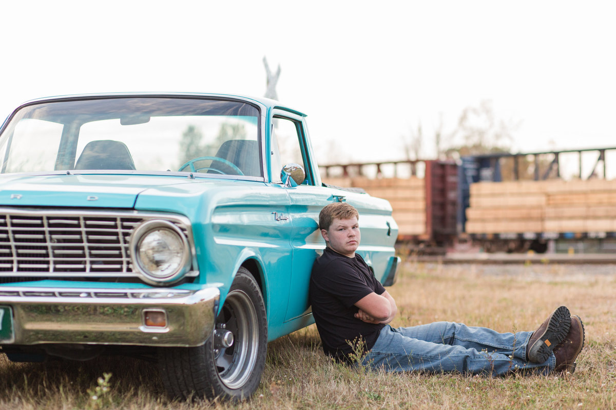 waynedale high school senior guy sitting on ground leaning against an old blue car with train tracks in the distance, photographed by Jamie Lynette Photography Canton Ohio Senior Photographer