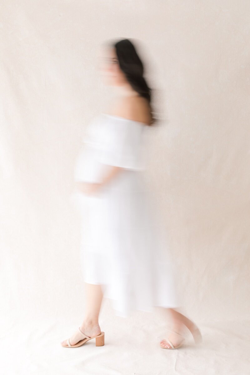 A pregnant woman wearing a white dress holds her stomach while walking across the frame. The motion of her walking is blurred.