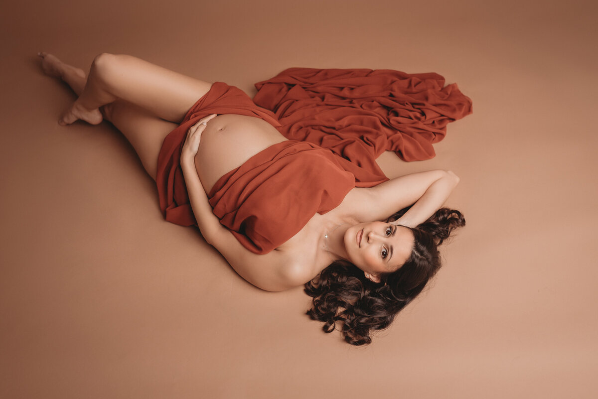 Pregnant woman laying on back draped in burnt orange chiffon fabric legs crossed with one hand on stomach and other hand behind her head