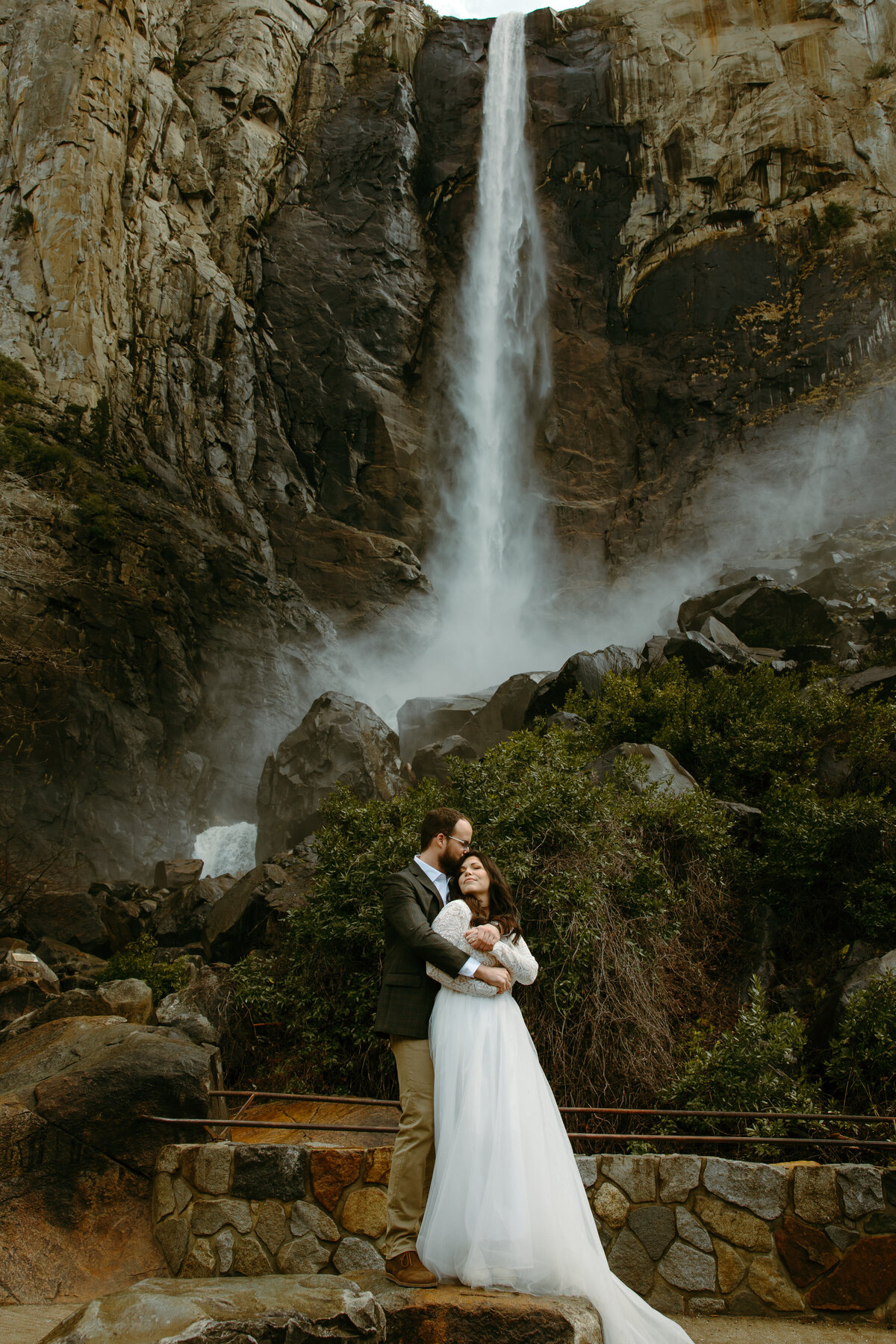 Bride and groom pose infront of a beautiful waterfall. Groom kissing the top of the brides head while she cuddles close to him.