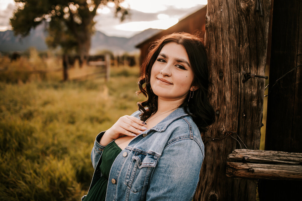 girl sits against a fence with a barn behind her for senior photos