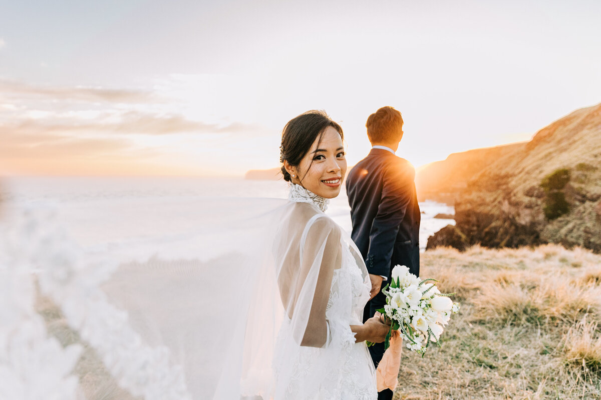 The groom leading the bride  towards the sunset on a cliff while the bride loos back into the camera and smile.