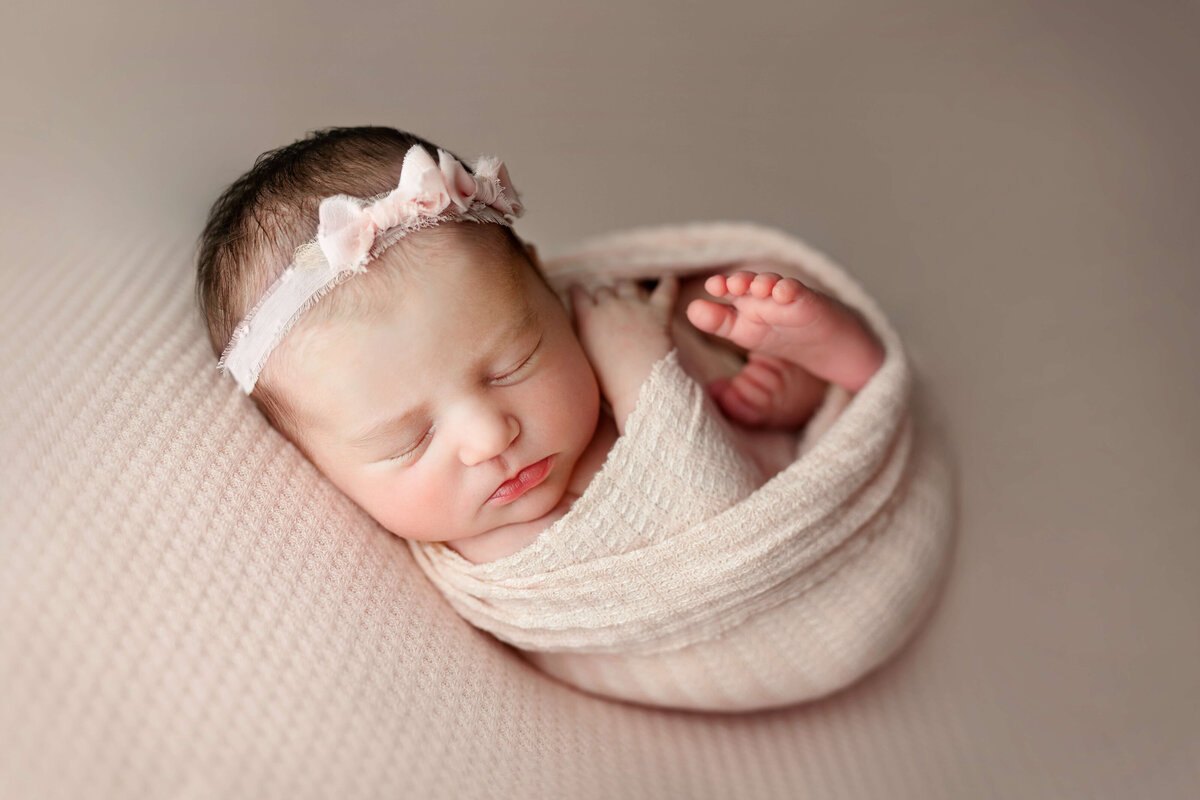 newborn baby girl wrapped in a light pink wrap and pink headband with bows laying on a matching textured backdrop at a loudoun county va newborn photo session