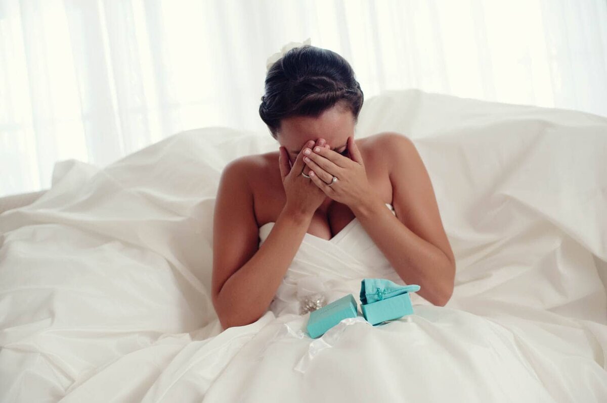 a bride surrounded by her white dress holds a blue tiffany's box and covers her face as she cries