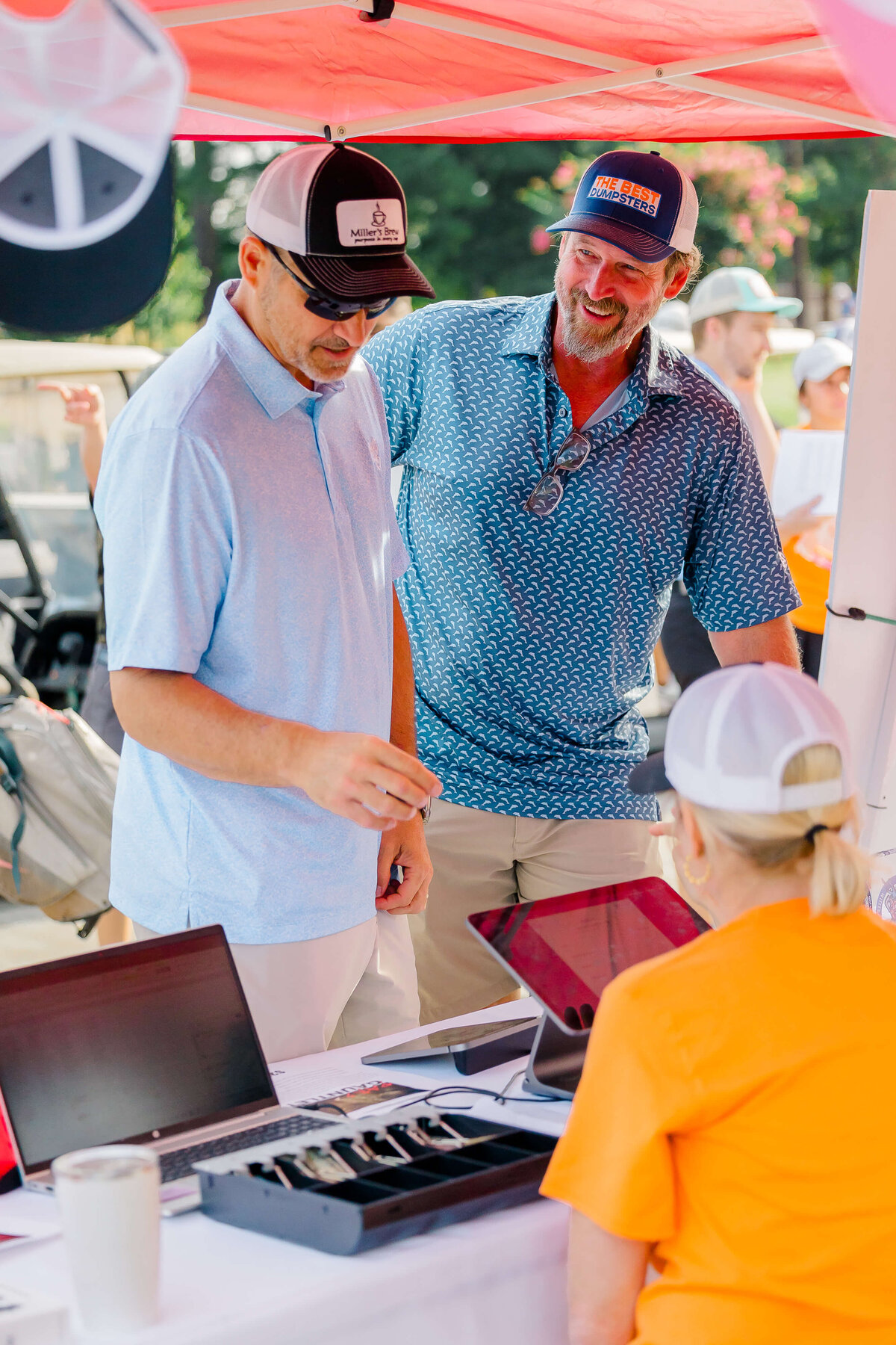 Two men wearing hats stand at a booth with laptops, chatting to a seated person under a red canopy. The men, dressed in casual shirts and shorts, engage with the person in an orange shirt and white hat. An event photographer captures the lively scene in Fayetteville, NC, with people bustling in the background.