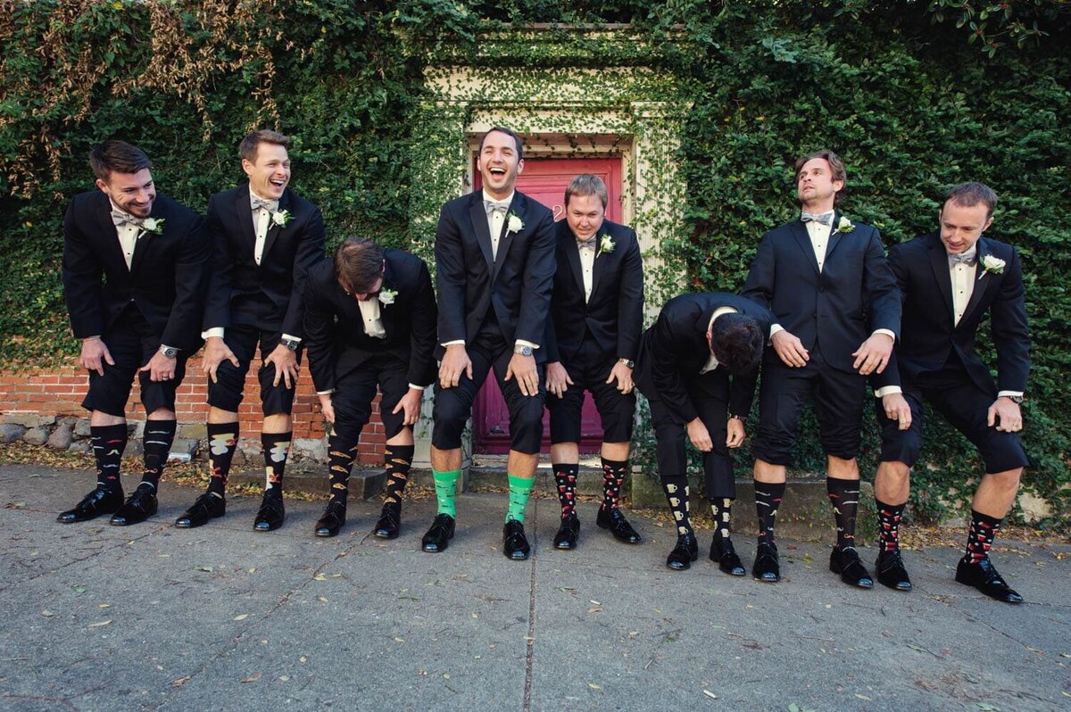 a groom and 7 groomsmen laugh as they  pull up their pant legs to show off their funny socks