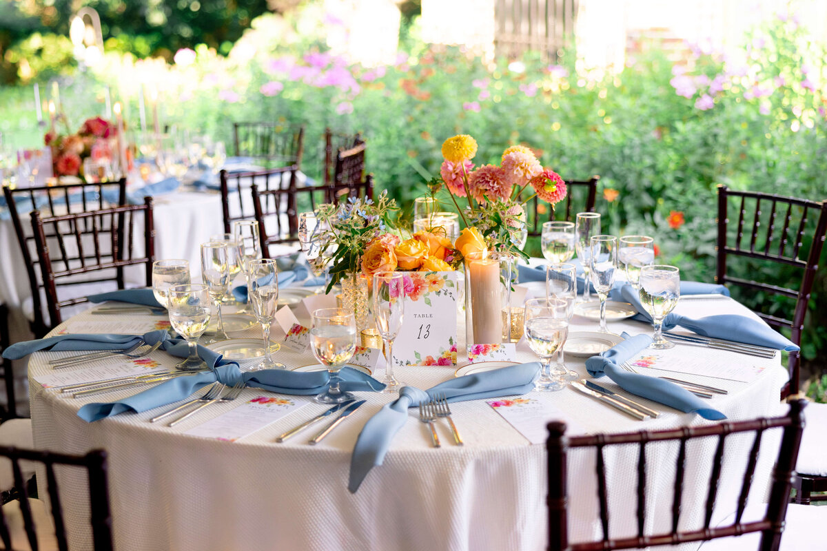 A beautifully set round table outdoors with elegant glassware, white napkins folded with blue ribbon, and a floral centerpiece featuring pink and orange flowers. A numbered table card is displayed in the center. Garden greenery is visible in the background.