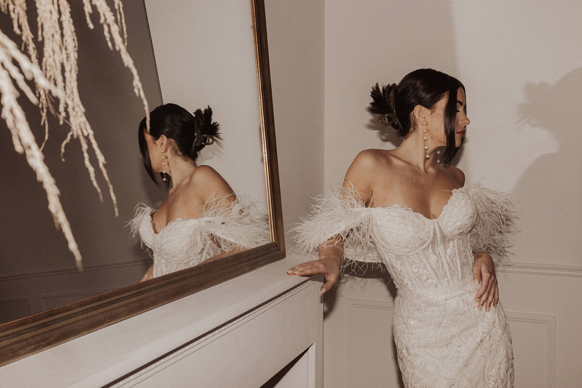 A bride in an off-shoulder gown with feathers, reflected in a mirror.