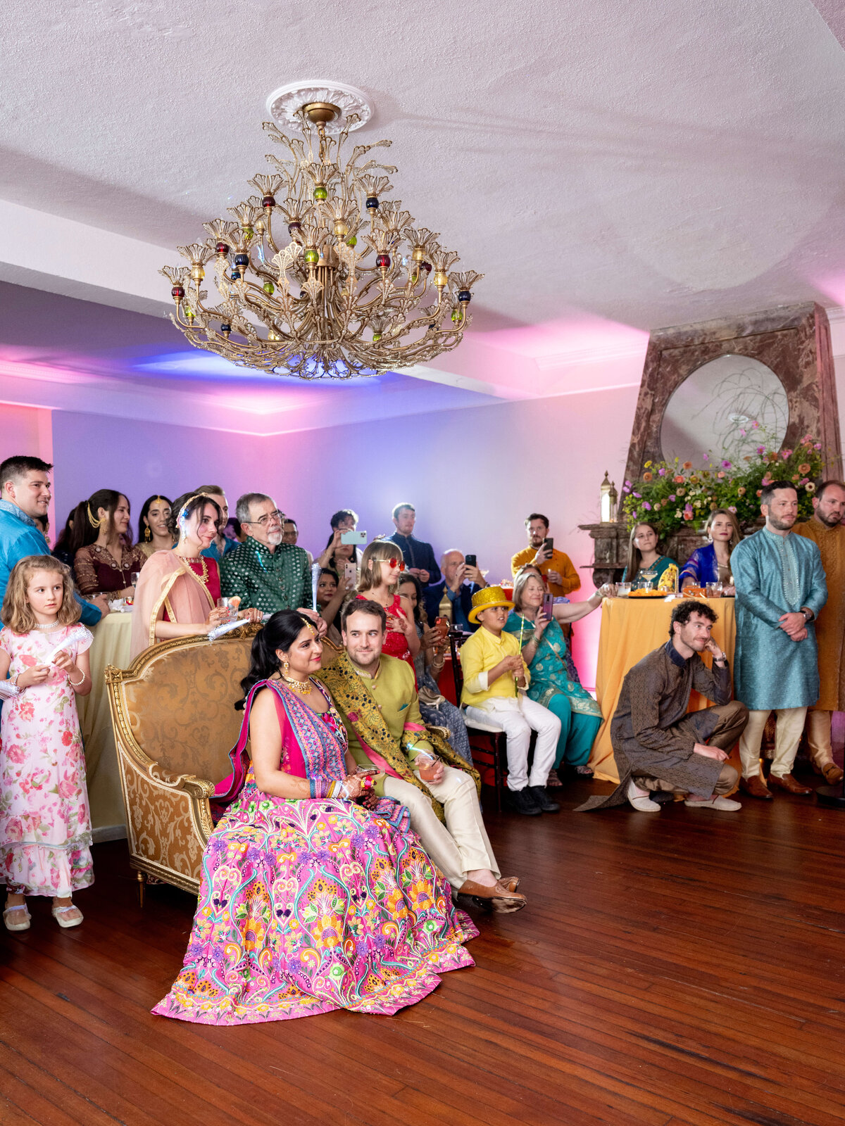 A vibrant group of people dressed in colorful traditional attire gather in a decorated room. They sit and stand, looking forward. The room features a chandelier, floral decorations, and warm lighting, creating a festive atmosphere.