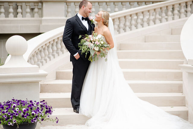 A newly married couple are smiling at each other while standing next to a white staircase