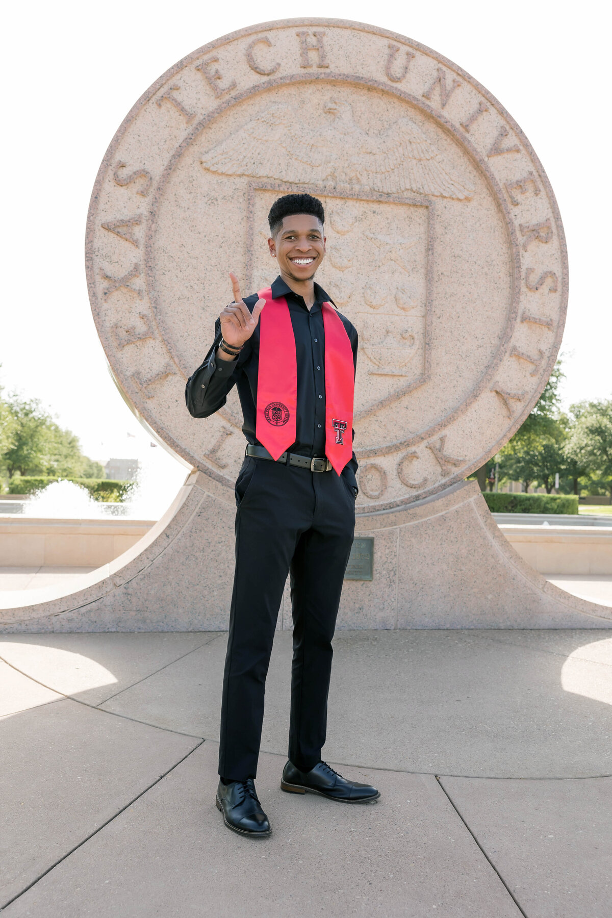 African  American male college senior photo session  on TTU Campus.  Male is wearing a black button down dress shirt with black pants and a red college stole and giving the  guns up while smiling at the camera