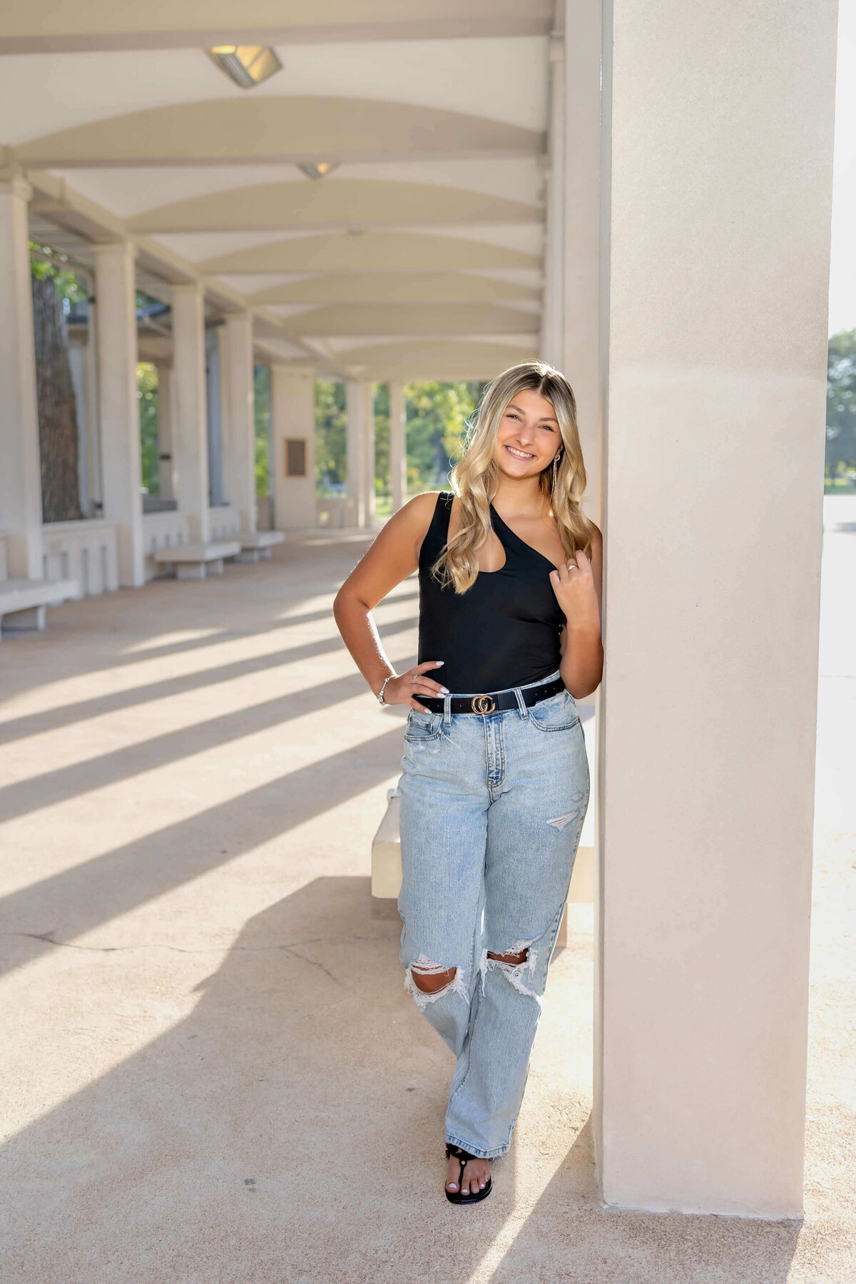 a blonde haired high school senior girl leaned against a concrete pillar of a covered walkway  outdoors full length image