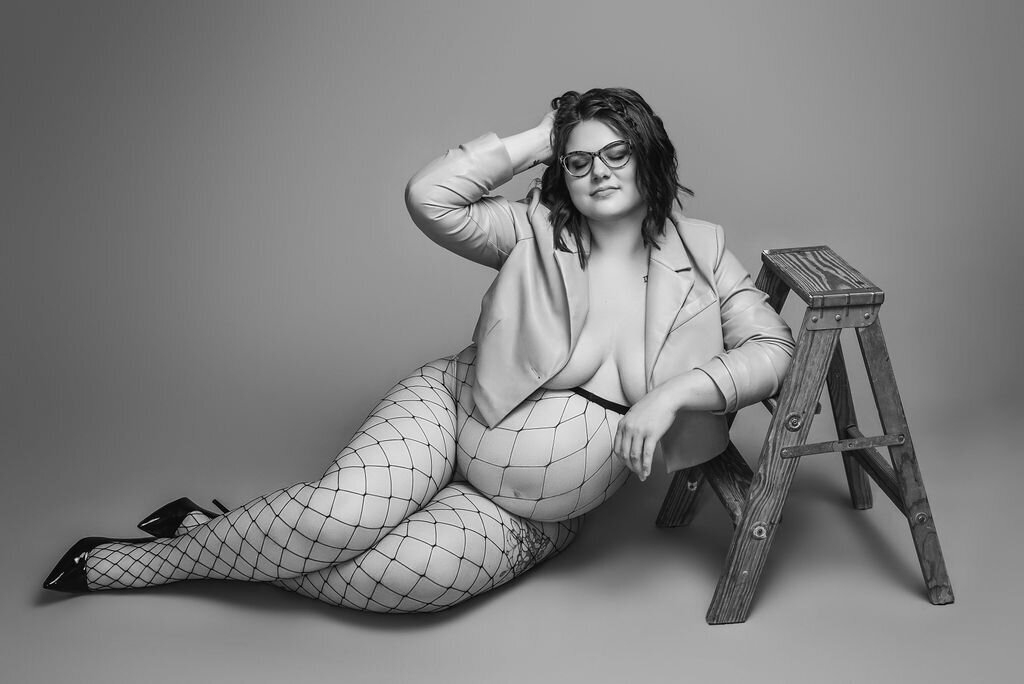 A pregnant woman in a light leather jacket, fishnets, and heels sits on the floor leaning against a wooden ladder in front of a neutral solid backdrop, captured by a photographer in Bemidji, Minnesota