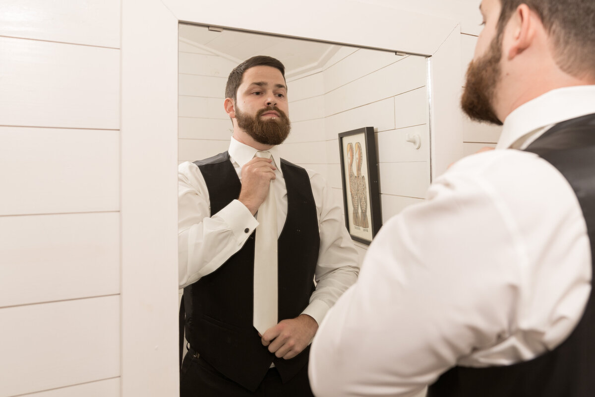 The groom ties his tie in the mirror as he gets ready for the wedding day.