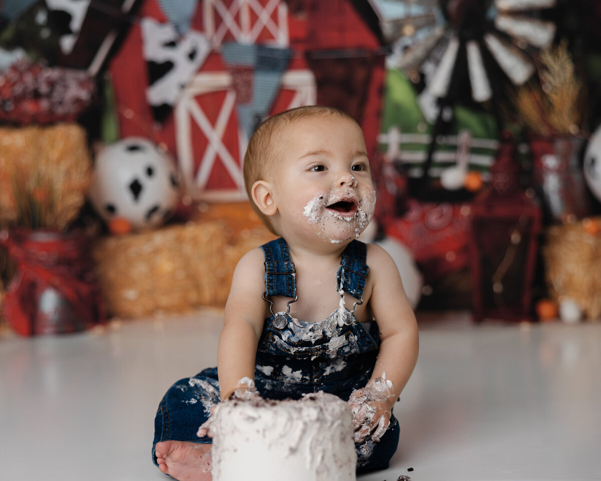 First birthday cake smash featuring a baby boy grinning while covered in cake and frosting, with a barn and hay scene behind him. Photograph by Yucaipa cake smash photographer Kaitlyn Dawn Photography.