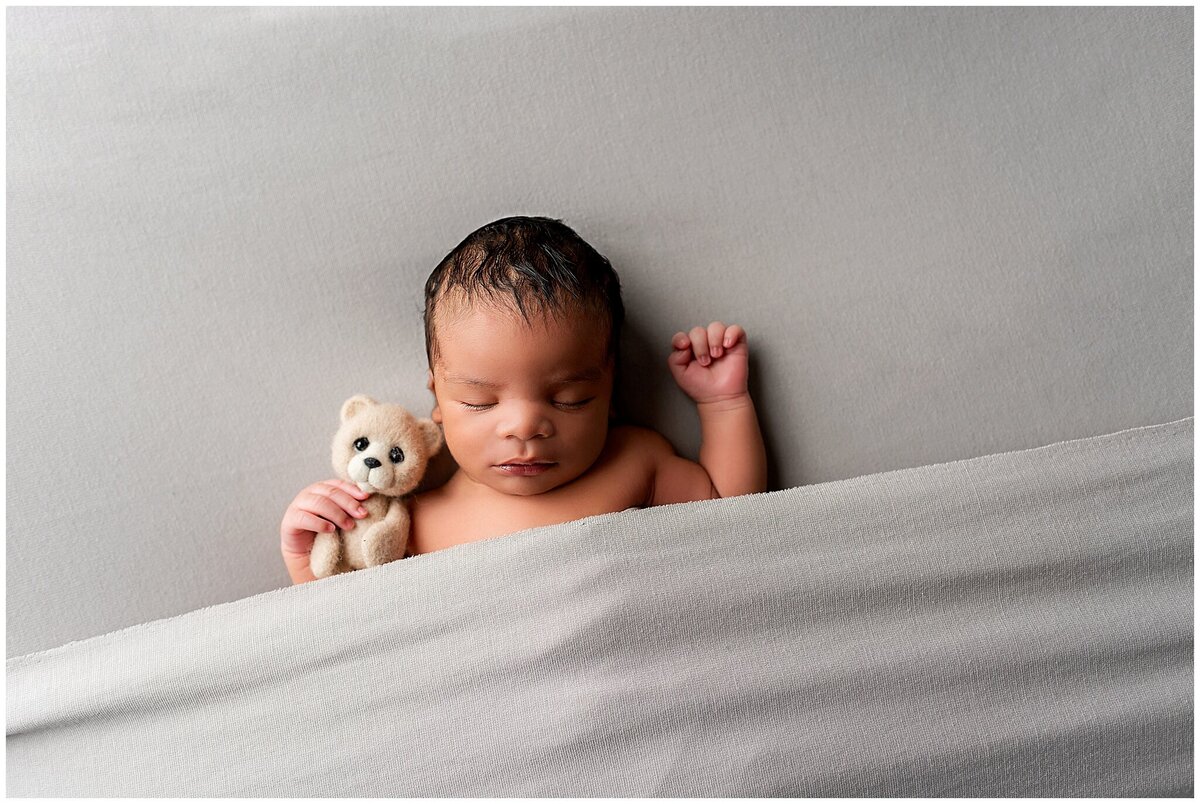 Newborn baby boy laying on his back with one arm up and the other arm holding a teddy bear.