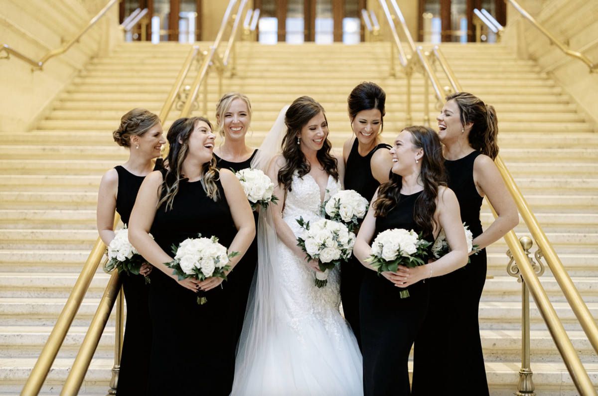 Timeless bride holds bouquet of white roses and smiles with six bridesmaids in black gowns stand on stairs at Harold Washington Library wedding reception.