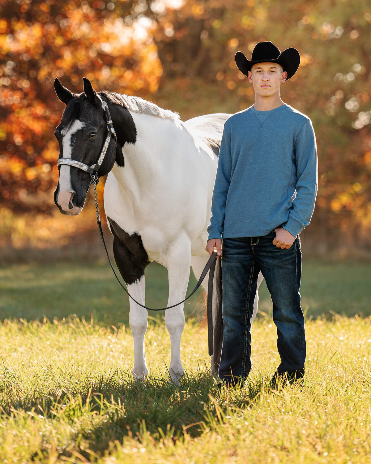male equestrian wearing a long sleeve blue shirt, jeans, and a black cowboy had standing proudly next to his black and white tobiano Paint horse gelding while looking into the camera with beautiful colored fall leaves behind them.