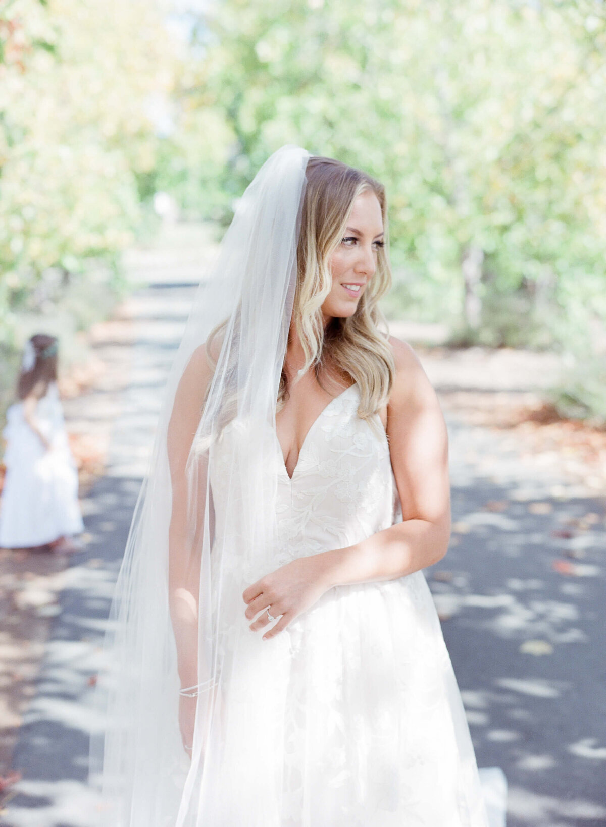 Beautiful bride with blond hair poses for the camera in her wedding ring and a veil on her head. Warm radiating greenery in the background.