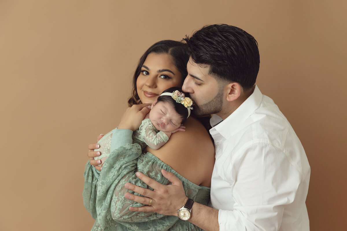 Happy new parents stand in a studio kissing their sleeping newborn daughter on mom's shoulder