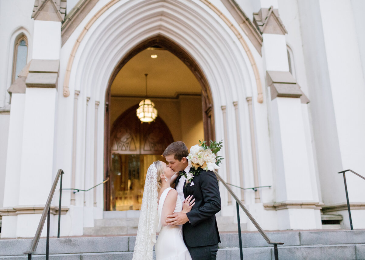 Couple kissing on the steps of Cathedral Basilica of St. John the Baptist with flowers around grooms neck.