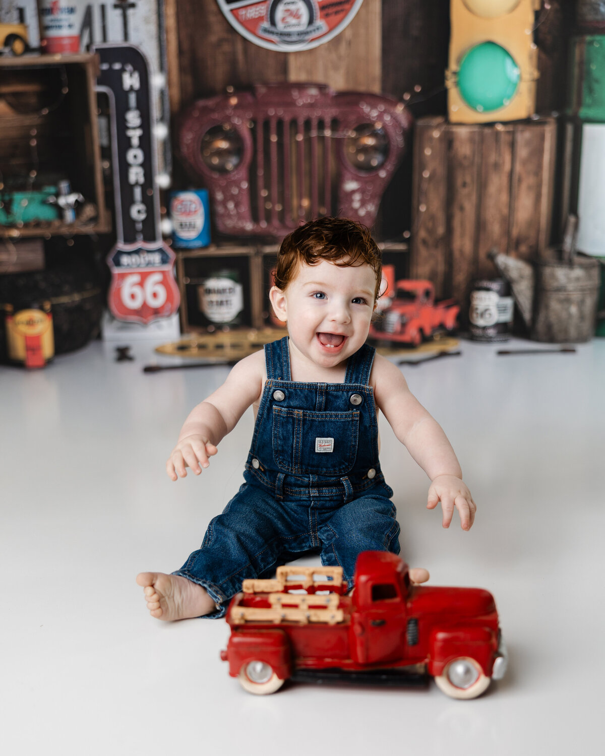 Smiling baby boy in denim overalls playing with a red toy truck in a garage-themed cake smash setup. Photograph by Yucaipa cake smash photographer Kaitlyn Dawn Photography.
