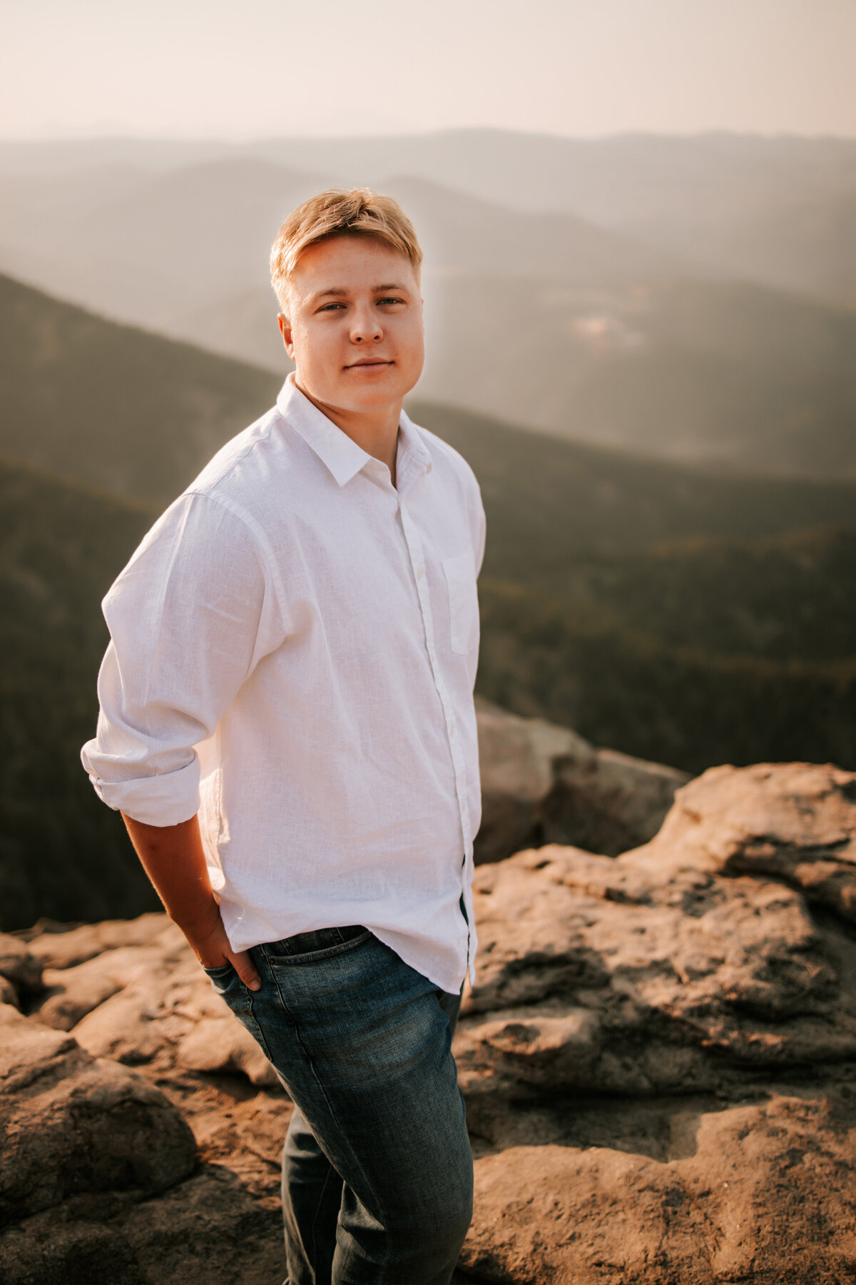 boy stands on top of mountain in boulder colorado for senior session in a white shirt during golden hour