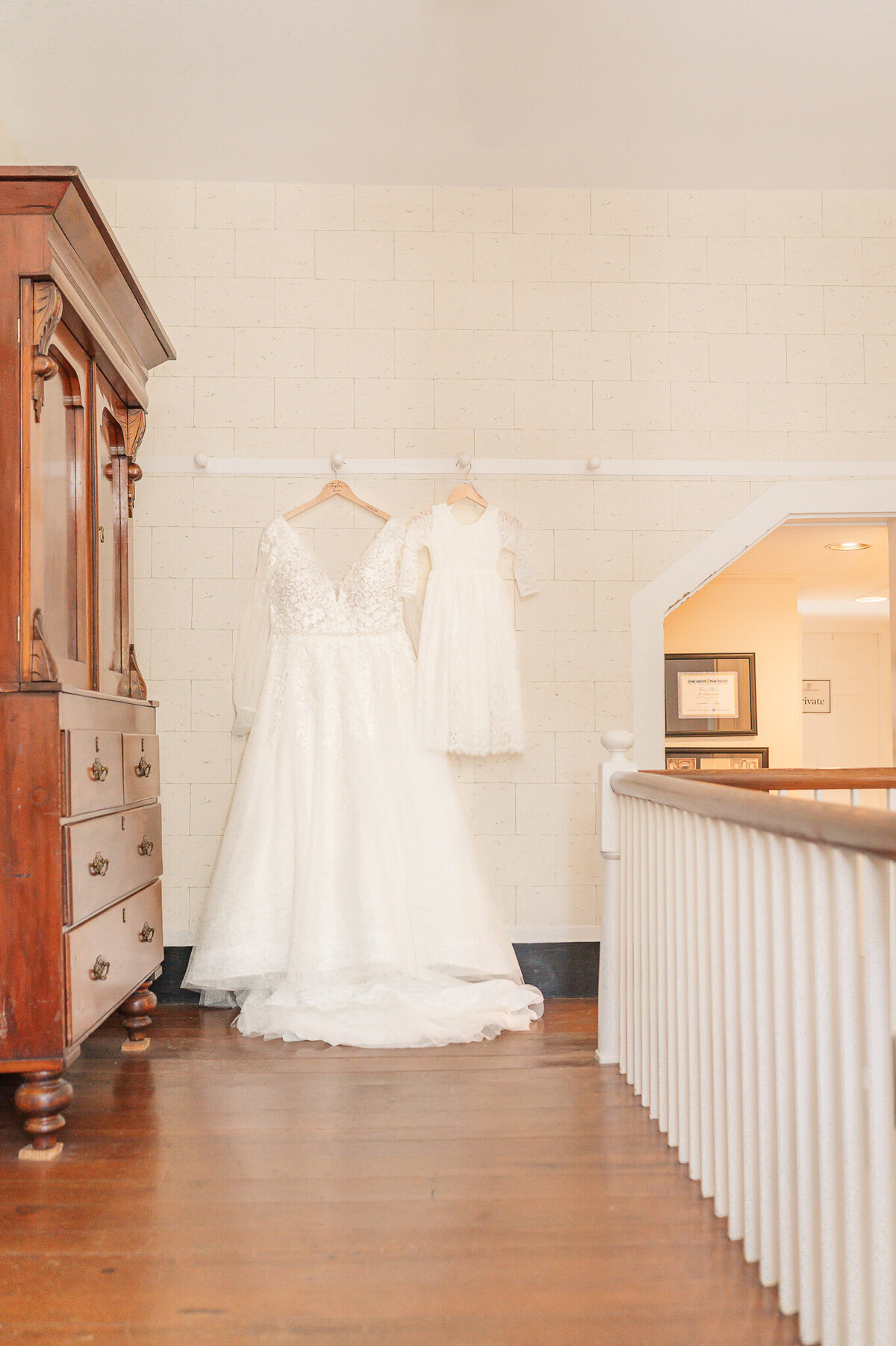 A wedding dress hanging with a flower girl's dress hanging in a hallway of The Sutherland by JoLynn Photography