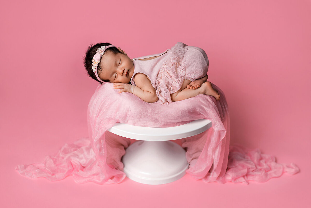 Newborn baby girl in pink romper posed on a cake stand.
