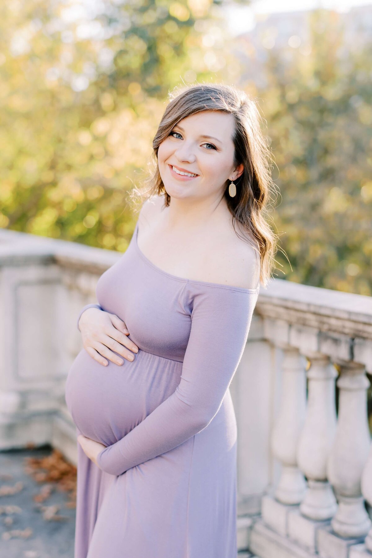 An expectant mom stands in front of a stately stone railing while cradling her growing bump.