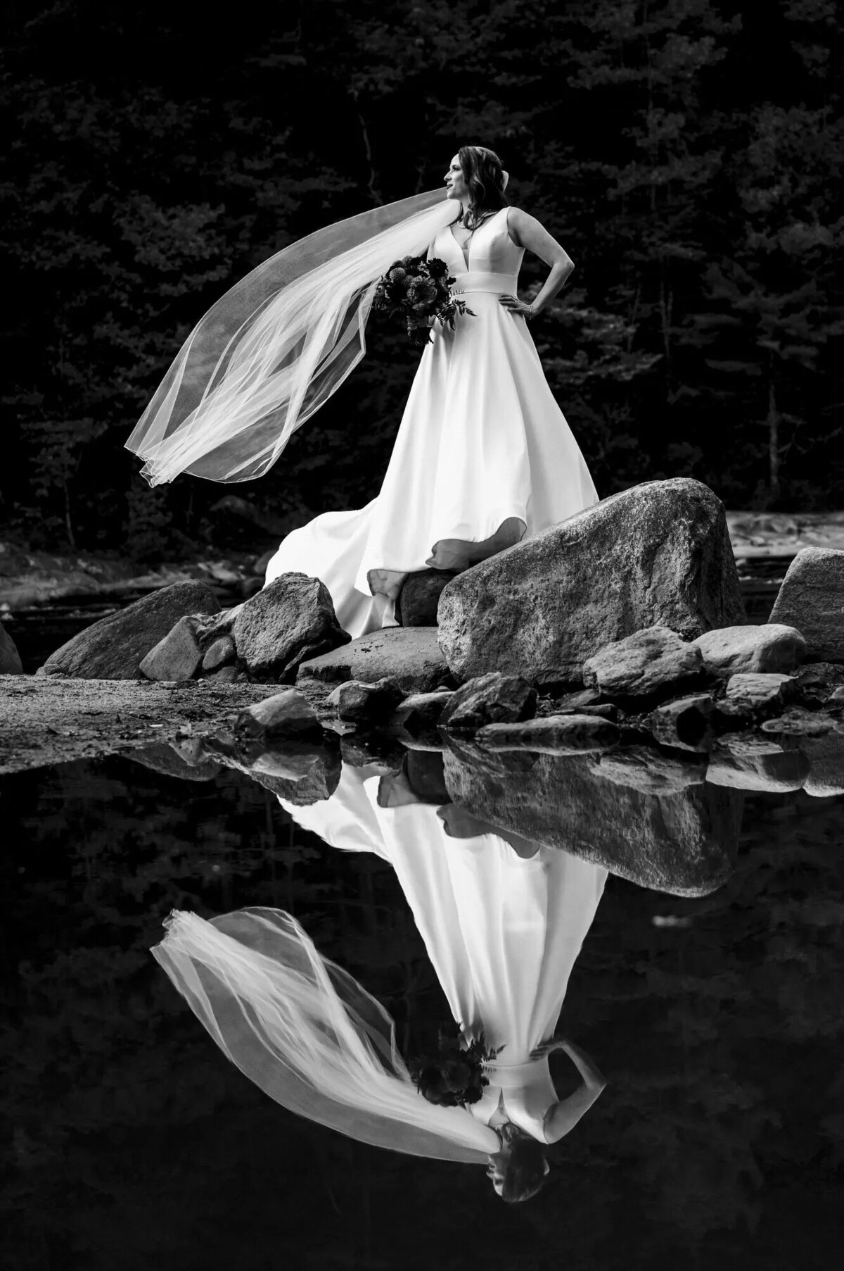 A stunning black and white image of a bride standing on a rock by a calm lake, her reflection and the elegant lines of her dress mirrored perfectly in the water.